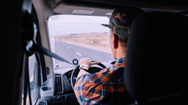 A look from behind or over the shoulder shot of a semi-truck driver in his cab as he looks onto the open road of a desert highway.