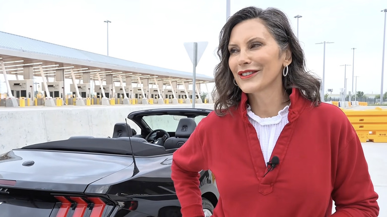 Michigan Governor Michigan Governor Gretchen Whitmer wearing a red shirt standing in front of a black Ford Mustang. She's smiling and looking off to her right